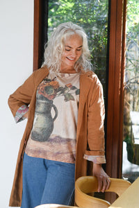 Woman wearing a floral t-shirt and brown coat standing indoors with a window in the background