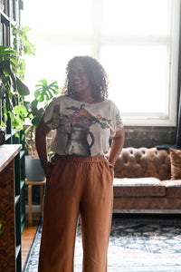 Woman standing in a living room wearing a floral top and brown pants.