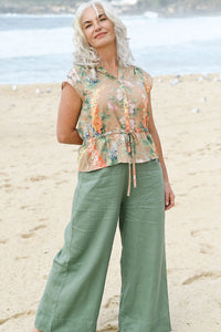 Woman wearing a floral top and green pants on a beach.