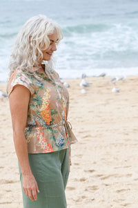 Woman standing on a beach wearing a floral sleeveless top and green pants.